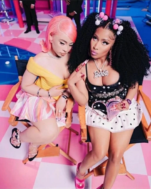 Two women, one with pink hair and the other with black hair and a Barbie necklace, pose in a photo studio with a checkered floor.