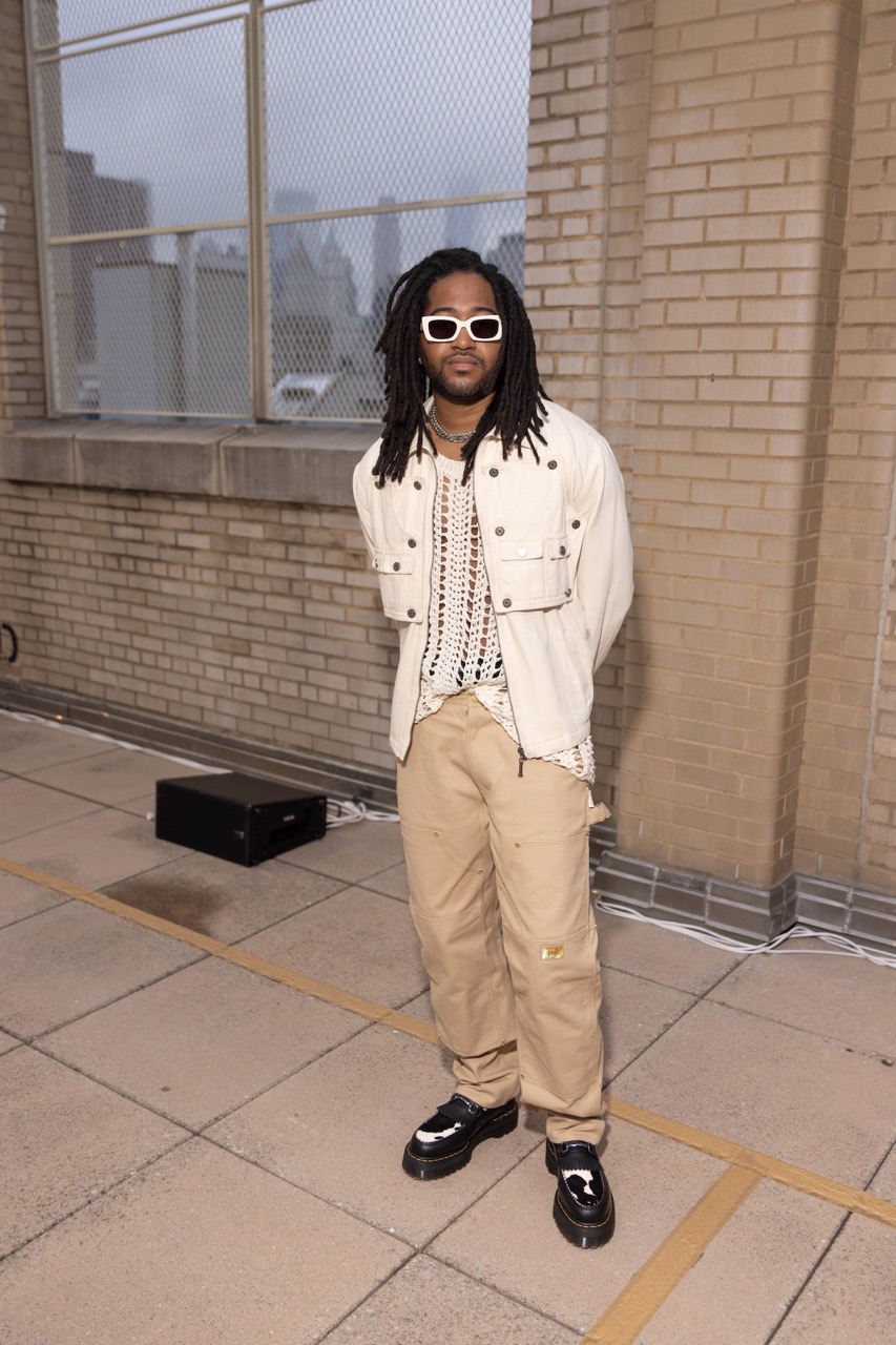 Man in streetwear style stands outdoors against a brick wall with urban background, wearing sunglasses, light jacket, and tan pants.