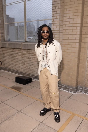 Man in streetwear style stands outdoors against a brick wall with urban background, wearing sunglasses, light jacket, and tan pants.