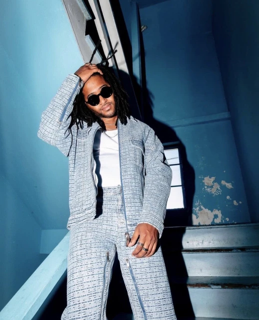 Stylish man with dreadlocks wearing sunglasses and a patterned suit poses indoors on a blue stairwell.