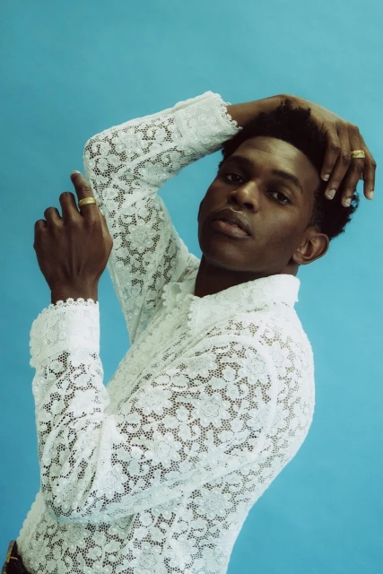 An androgynous person poses against a blue background, wearing a white lace shirt with long sleeves, hands above their head, looking at the camera.