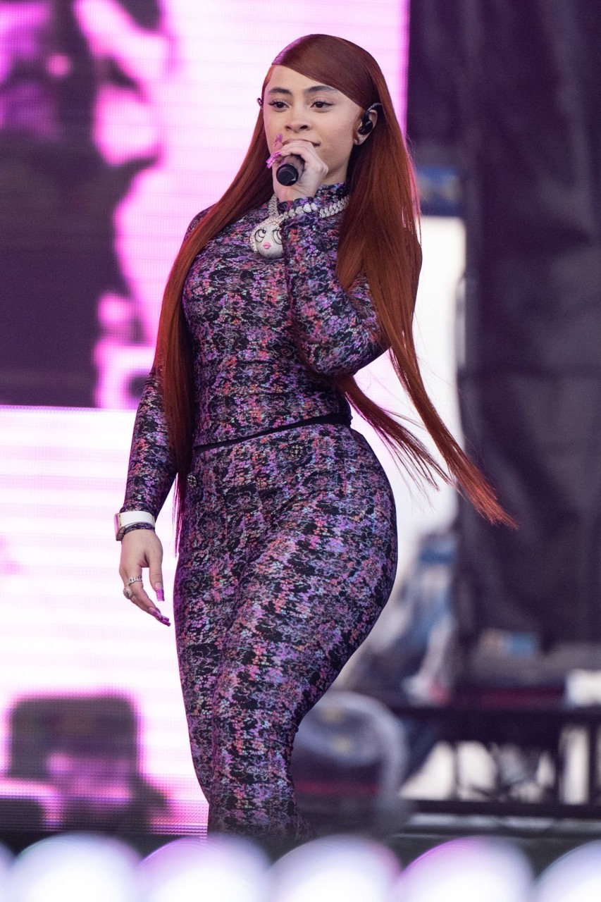Adult woman with long auburn hair performing on stage wearing a purple and black floral print jumpsuit and diamond necklace and bracelet.