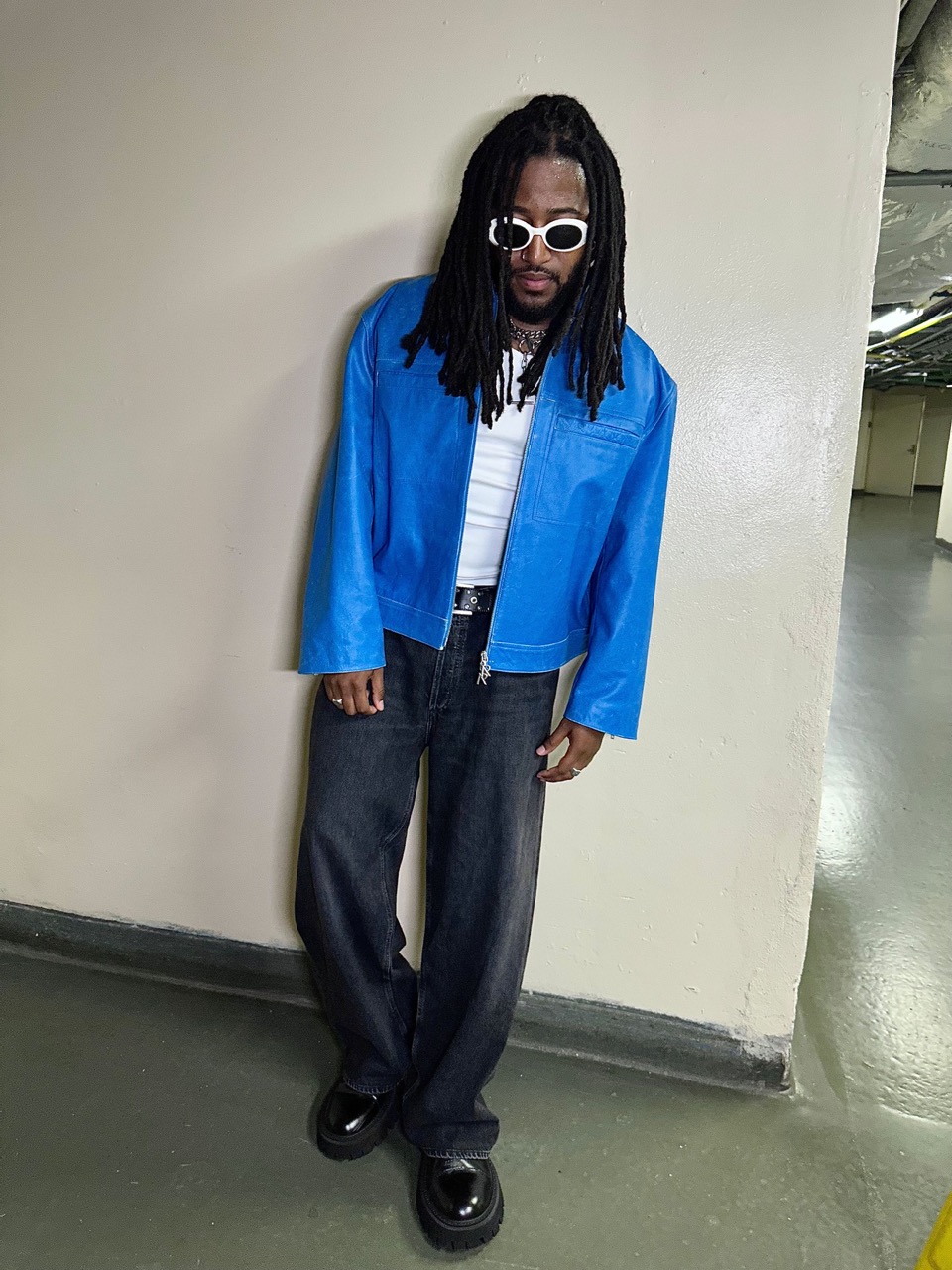 Adult male in blue jacket and black jeans poses against a neutral wall, wearing sunglasses and chunky-soled shoes in an indoor setting.