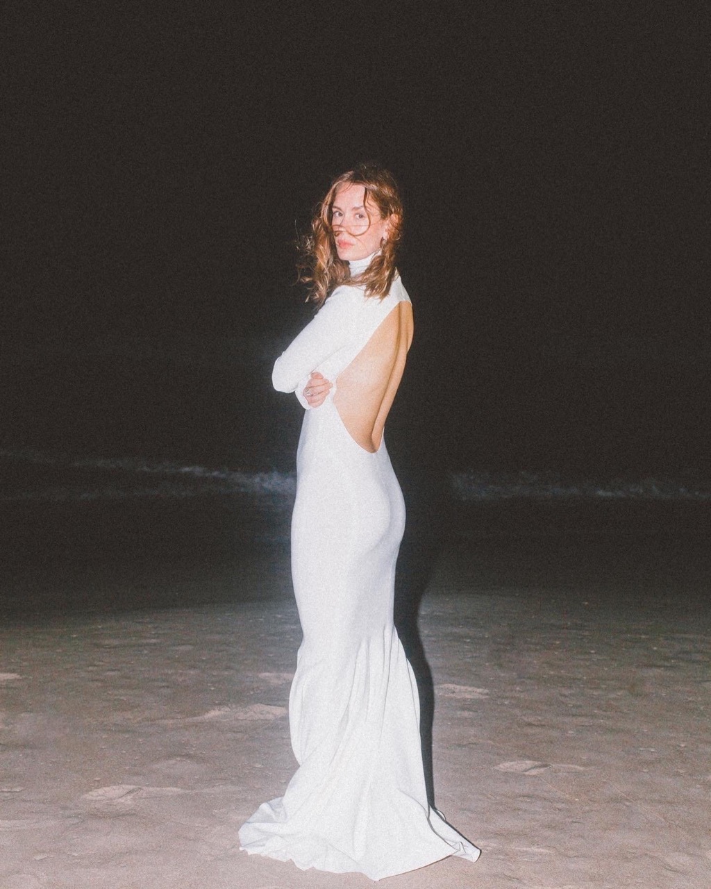 Woman in a white gown stands on a beach at night, arms crossed, with an open back, and ocean waves visible in the dark background.