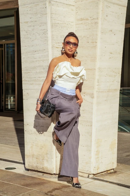 A fashionable adult female stands against a neutral stone wall, showcasing a strapless white top, wide-leg trousers, and stylish accessories.