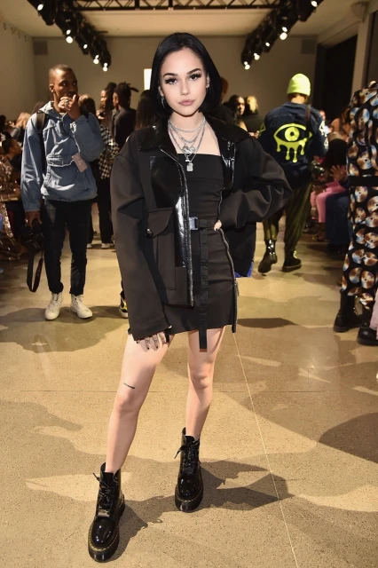Fashionable young woman in all-black outfit strikes a pose at an indoor fashion show, with a crowd of onlookers in the background.