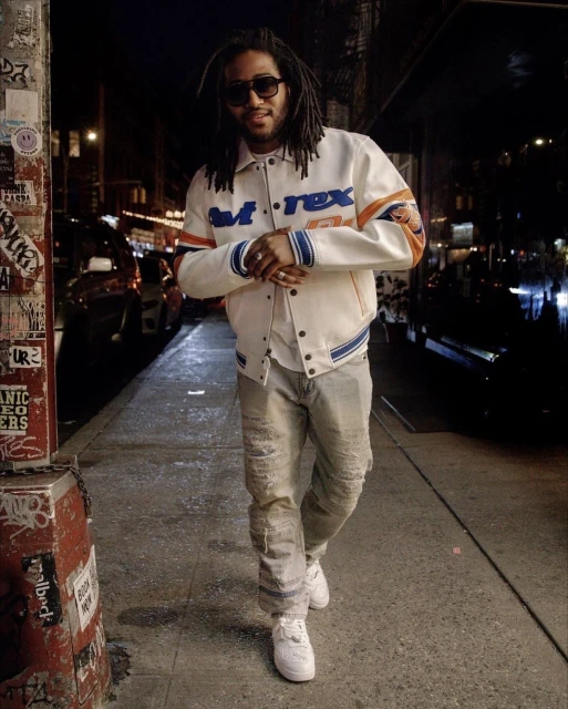 A man with locs and sunglasses walks on a city sidewalk at night wearing a letterman jacket and white sneakers.