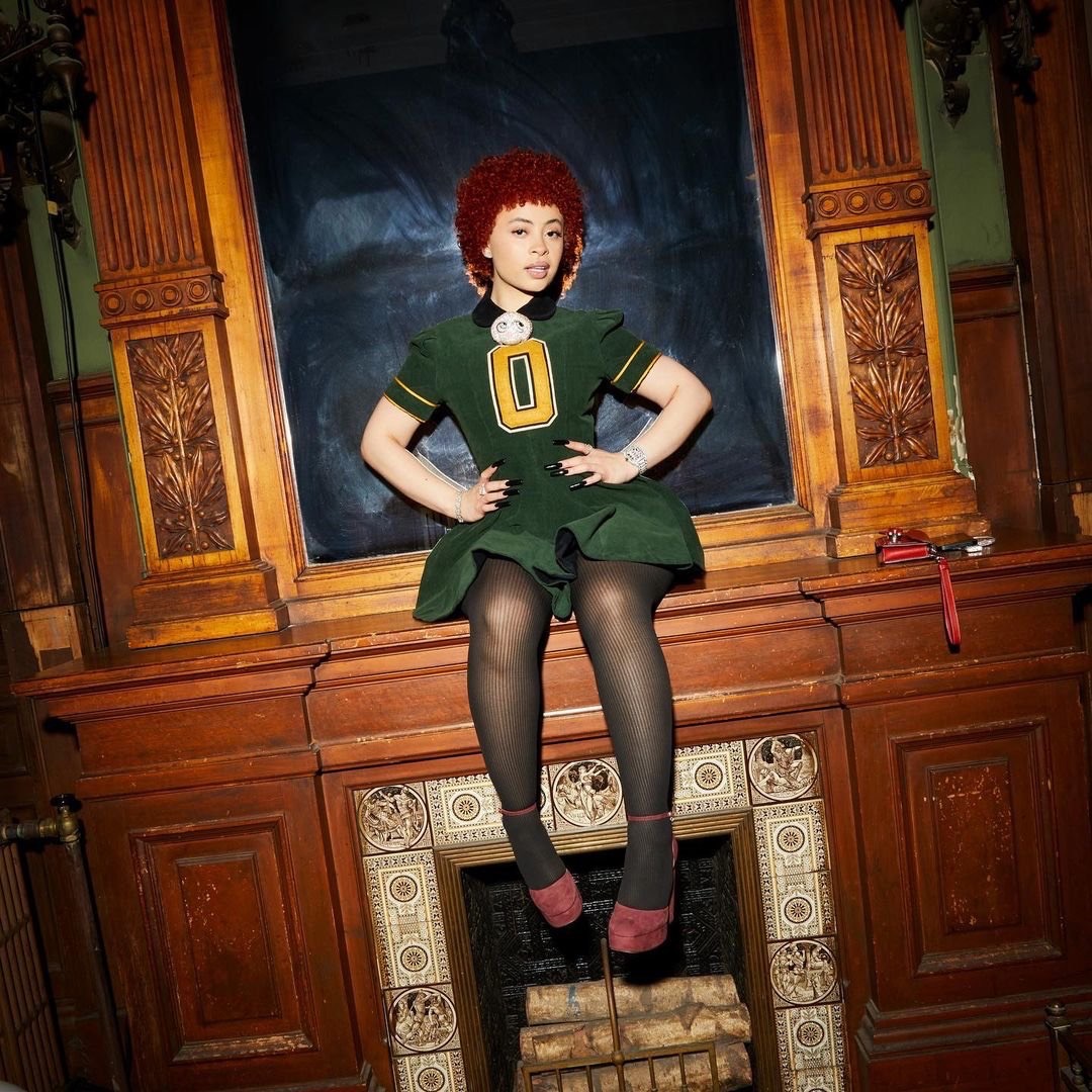 Ice Spice poses indoors, wearing a green dress, black tights, and red platform heels, seated on a decorative wooden fireplace mantel.