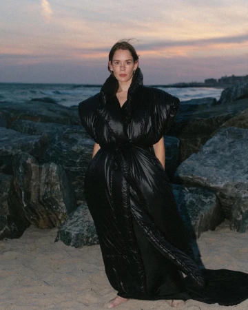 Woman poses on a beach wearing a black puffer coat. Ocean waves crash in the background during sunset. Large rocks are scattered around.