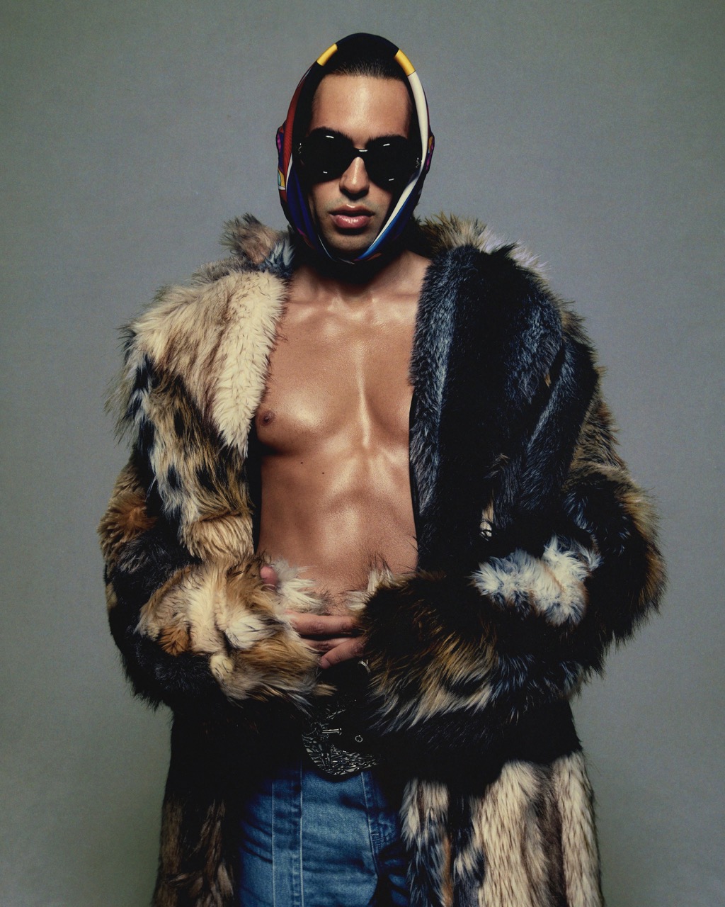 An adult male poses in a fur coat and jeans with a headscarf and sunglasses against a plain gray backdrop in a studio setting.