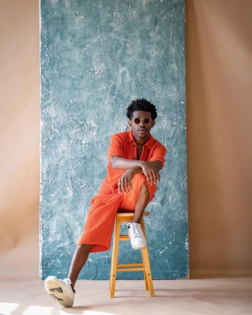 Adult male in orange outfit sits on a wooden stool in front of a textured blue panel in a studio setting. He wears round sunglasses.
