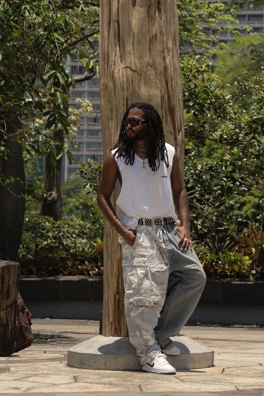 Adult male with dreadlocks wearing a white tank top and patchwork jeans leans against a tree trunk outdoors in an urban setting.