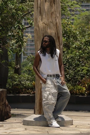 Adult male with dreadlocks wearing a white tank top and patchwork jeans leans against a tree trunk outdoors in an urban setting.