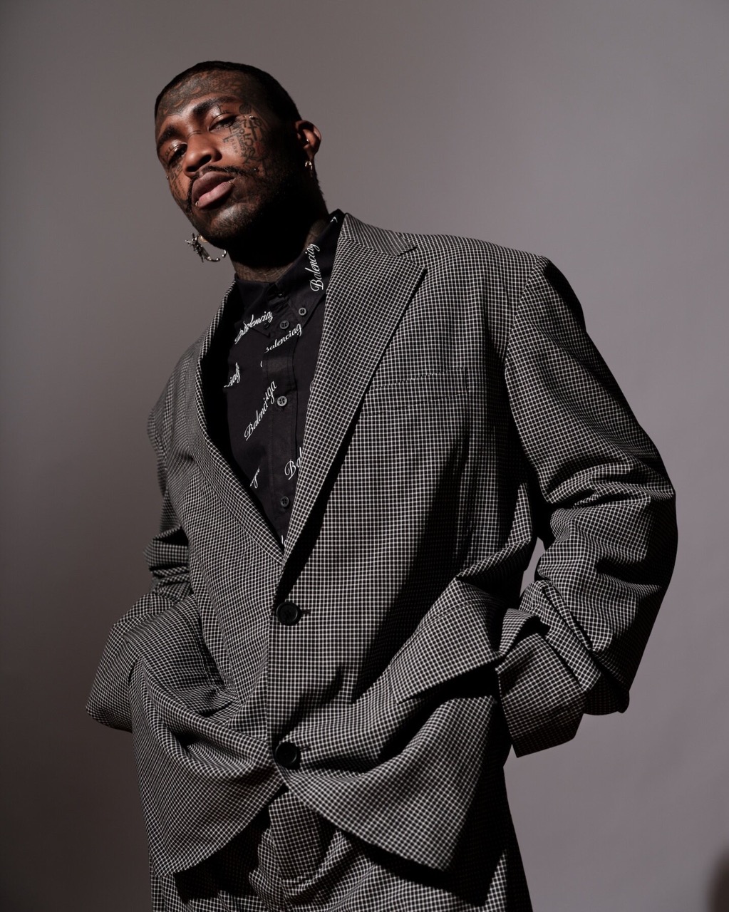 Portrait of a Black adult male in a gray houndstooth suit and patterned shirt, looking at the camera against a gray background.