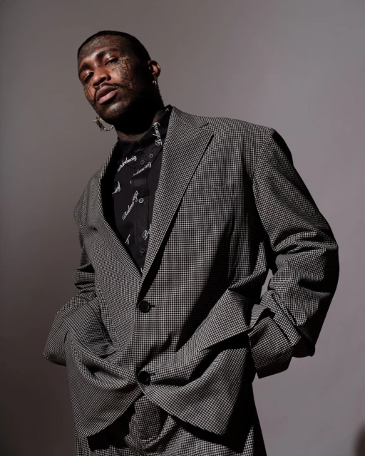 Portrait of a Black adult male in a gray houndstooth suit and patterned shirt, looking at the camera against a gray background.