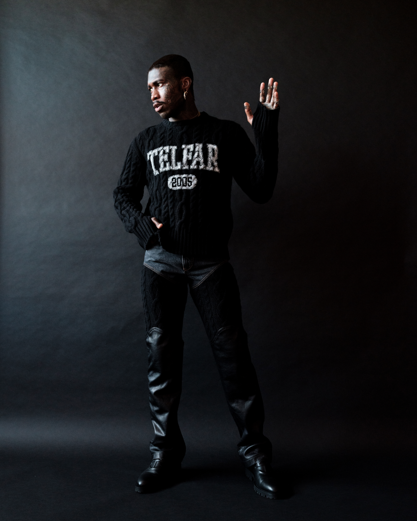 Man in a black Telfar sweater and black chaps poses in a studio. He has an arm raised and is looking to the left.