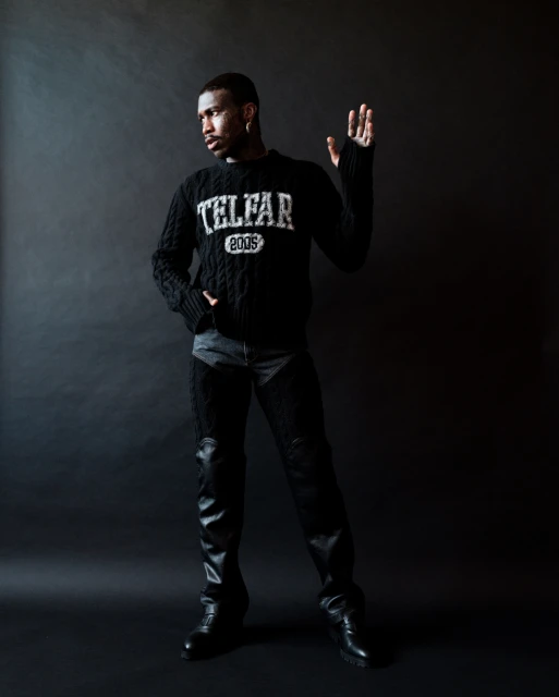 Man in a black Telfar sweater and black chaps poses in a studio. He has an arm raised and is looking to the left.