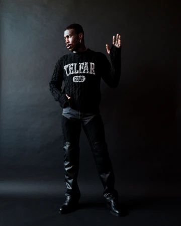 Man in a black Telfar sweater and black chaps poses in a studio. He has an arm raised and is looking to the left.