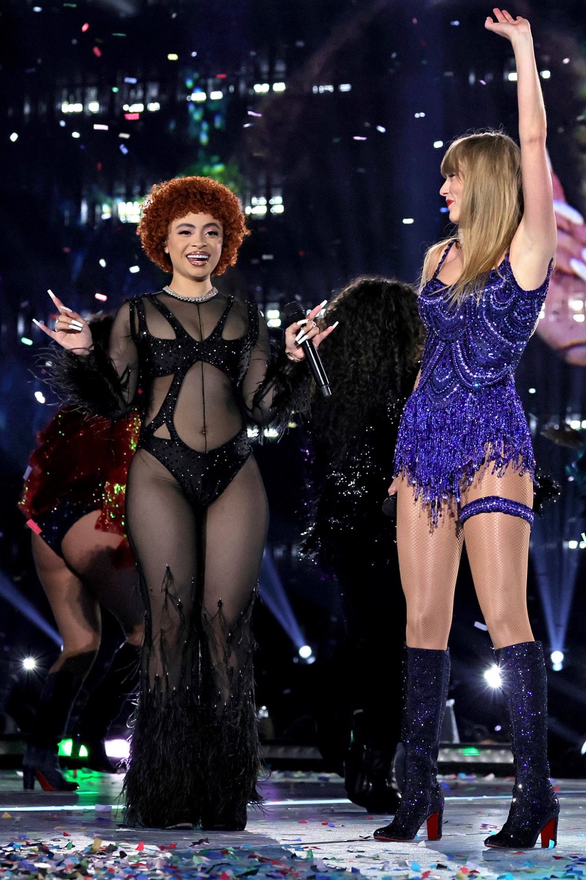 Two women, Ice Spice and Taylor Swift, stand on stage together in sparkly outfits, illuminated by stage lighting and confetti on the ground.