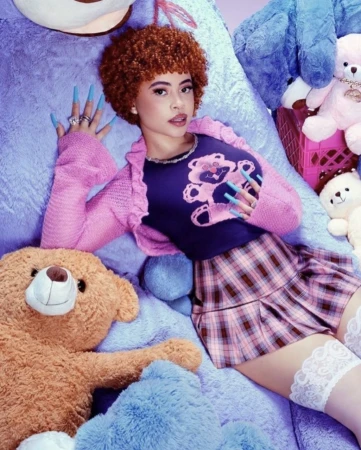 Adult female poses lying down with brown hair, wearing a pink cardigan and plaid skirt, surrounded by blue and brown teddy bears.