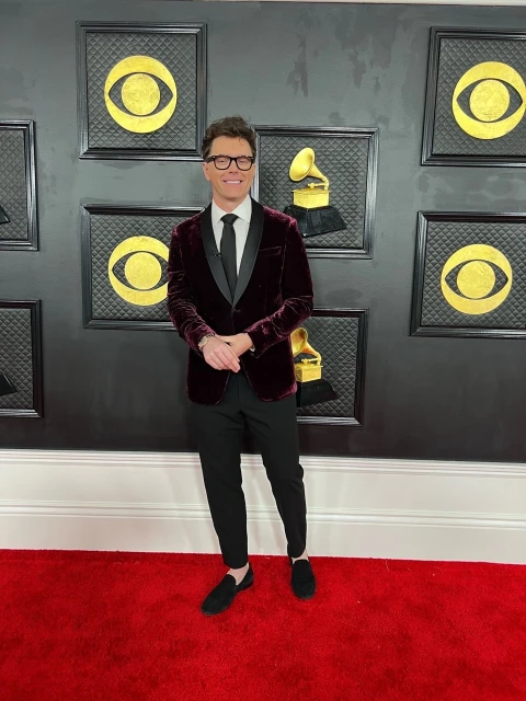 Adult male with eyeglasses poses on a red carpet wearing a velvet jacket and black pants with Grammy Awards background images.