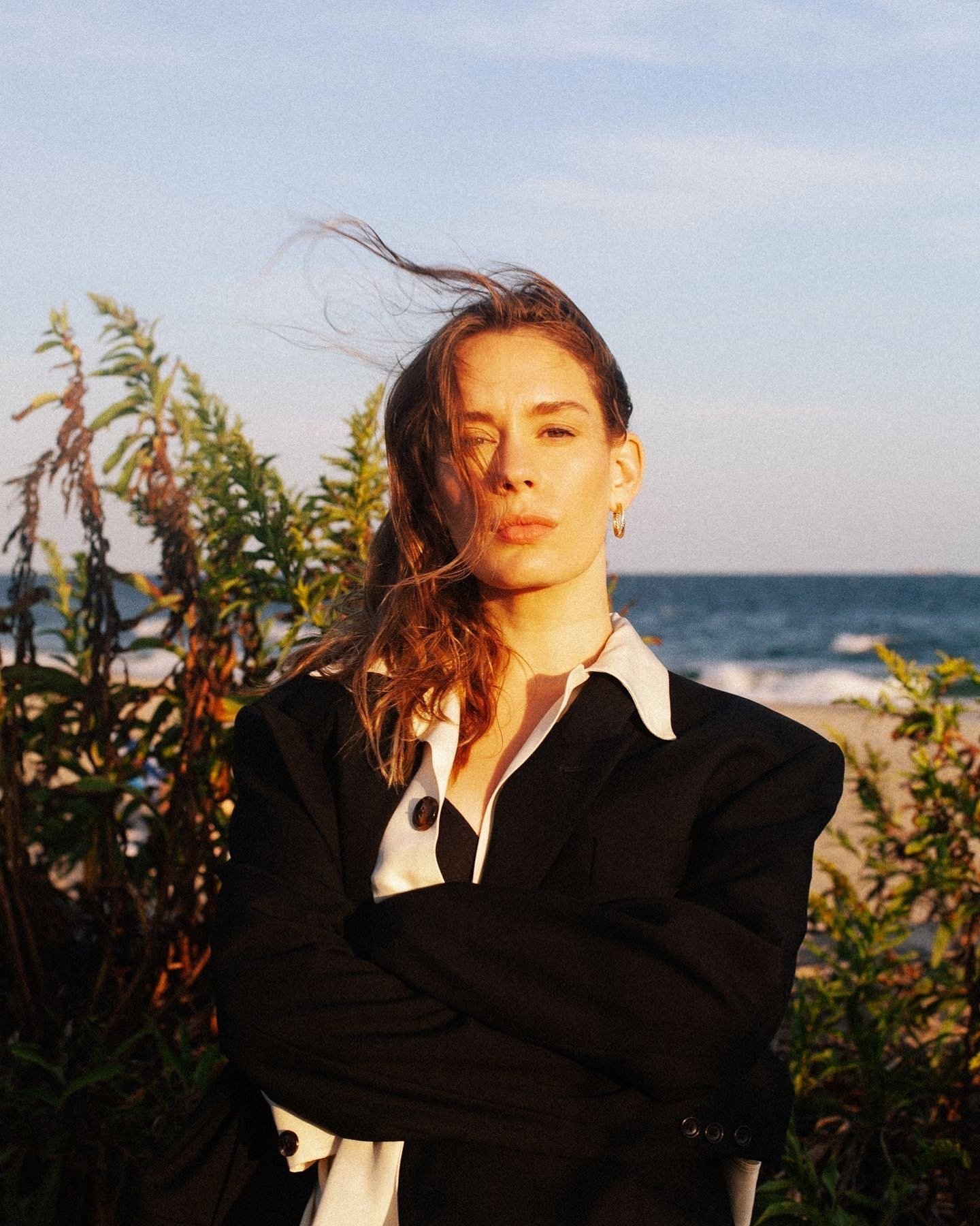 Woman in black blazer stands outside near beach on sunny day with foliage and blonde hair blowing in wind, eye-level, medium shot.