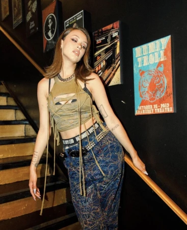 Young woman with medium brown hair poses on dark wooden stairs. She is wearing a brown tank top with cut-outs and patterned pants.