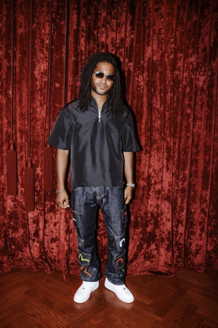 Man with dreadlocks, wearing sunglasses and a black shirt, stands in front of a red velvet curtain with a wood floor.