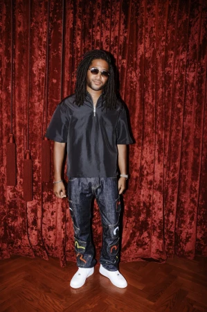 Man with dreadlocks, wearing sunglasses and a black shirt, stands in front of a red velvet curtain with a wood floor.