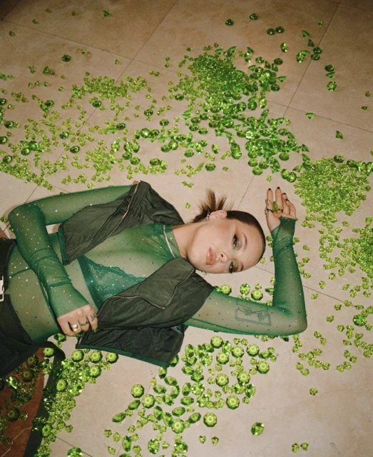 Woman lies on patterned floor amid green gemstones. She is wearing a sheer green top, black vest, and has a tattoo on her arm.
