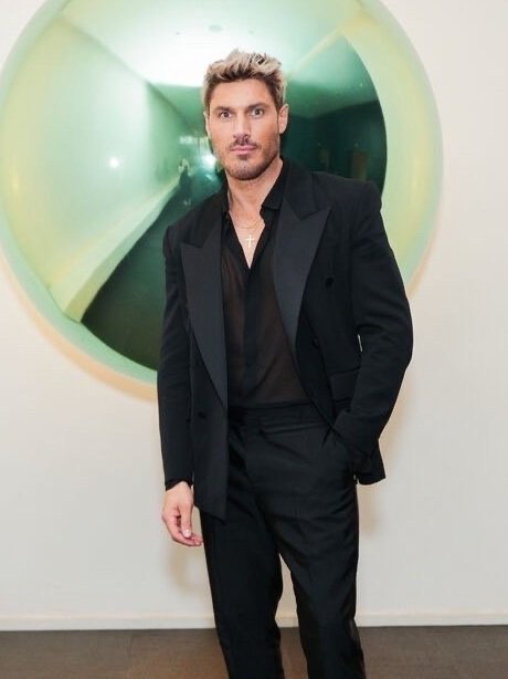 Man in a black suit poses in front of a silver balloon. He has blond hair, a sheer black shirt, and a gold necklace, indoors.