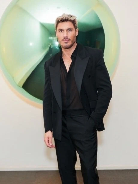 Man in a black suit poses in front of a silver balloon. He has blond hair, a sheer black shirt, and a gold necklace, indoors.
