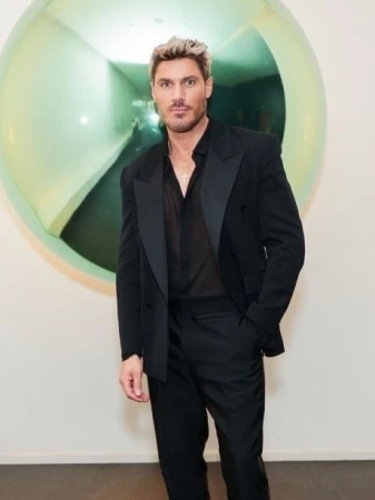 Man in a black suit poses in front of a silver balloon. He has blond hair, a sheer black shirt, and a gold necklace, indoors.