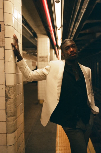 Fashion shot of an African-American man in a white blazer leaning against tiled wall in a subway station, looking at the camera.