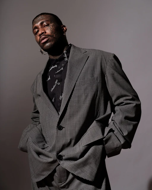 Portrait of a Black male in a grey suit, wearing a patterned shirt and earrings, facing the camera in a studio setting with grey backdrop.