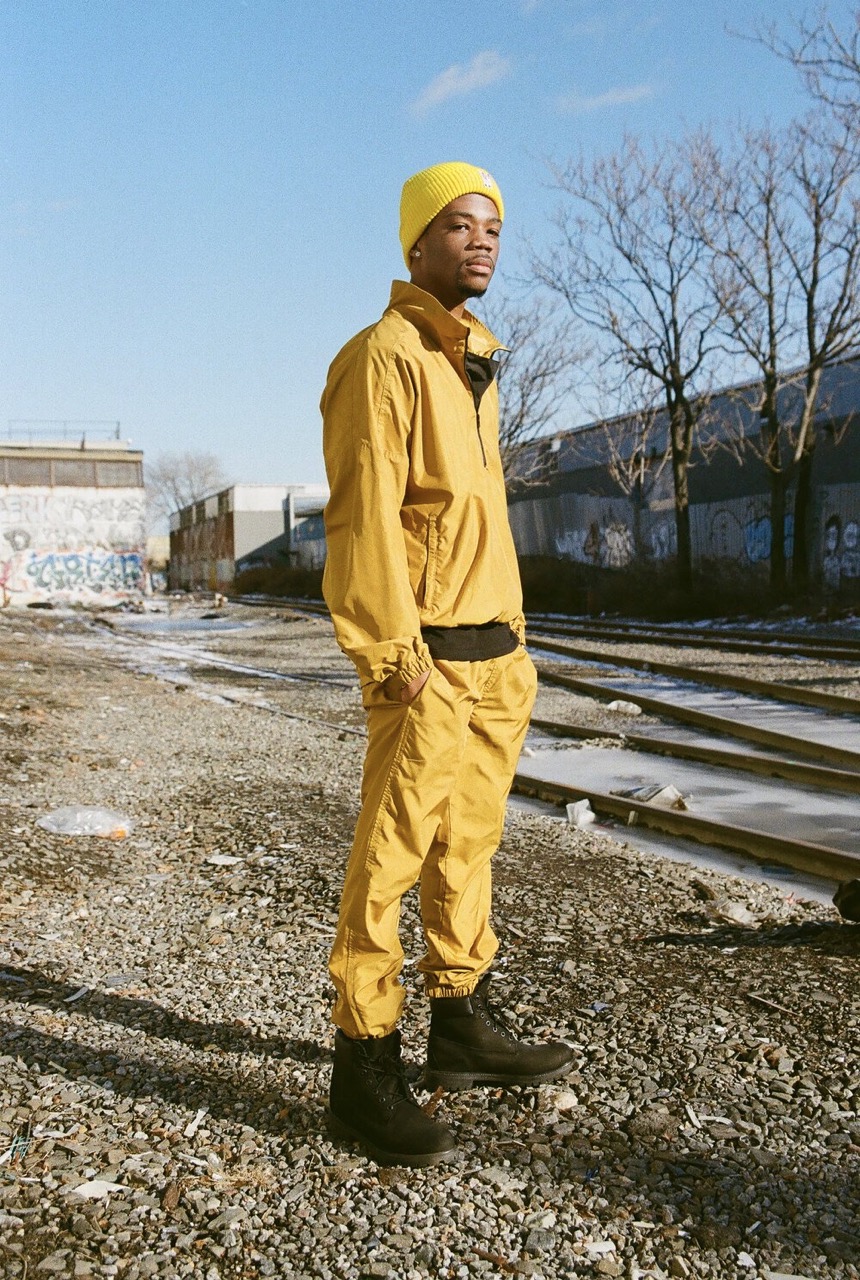 Young man in yellow tracksuit and beanie stands on gravel near train tracks against a sunny blue sky in an industrial area.