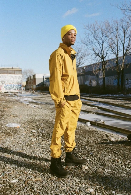 Young man in yellow tracksuit and beanie stands on gravel near train tracks against a sunny blue sky in an industrial area.