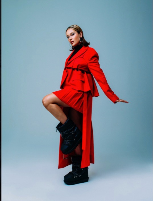 Woman poses in red skirt and jacket with black boots in a fashion shot against a light blue background in a photo studio.