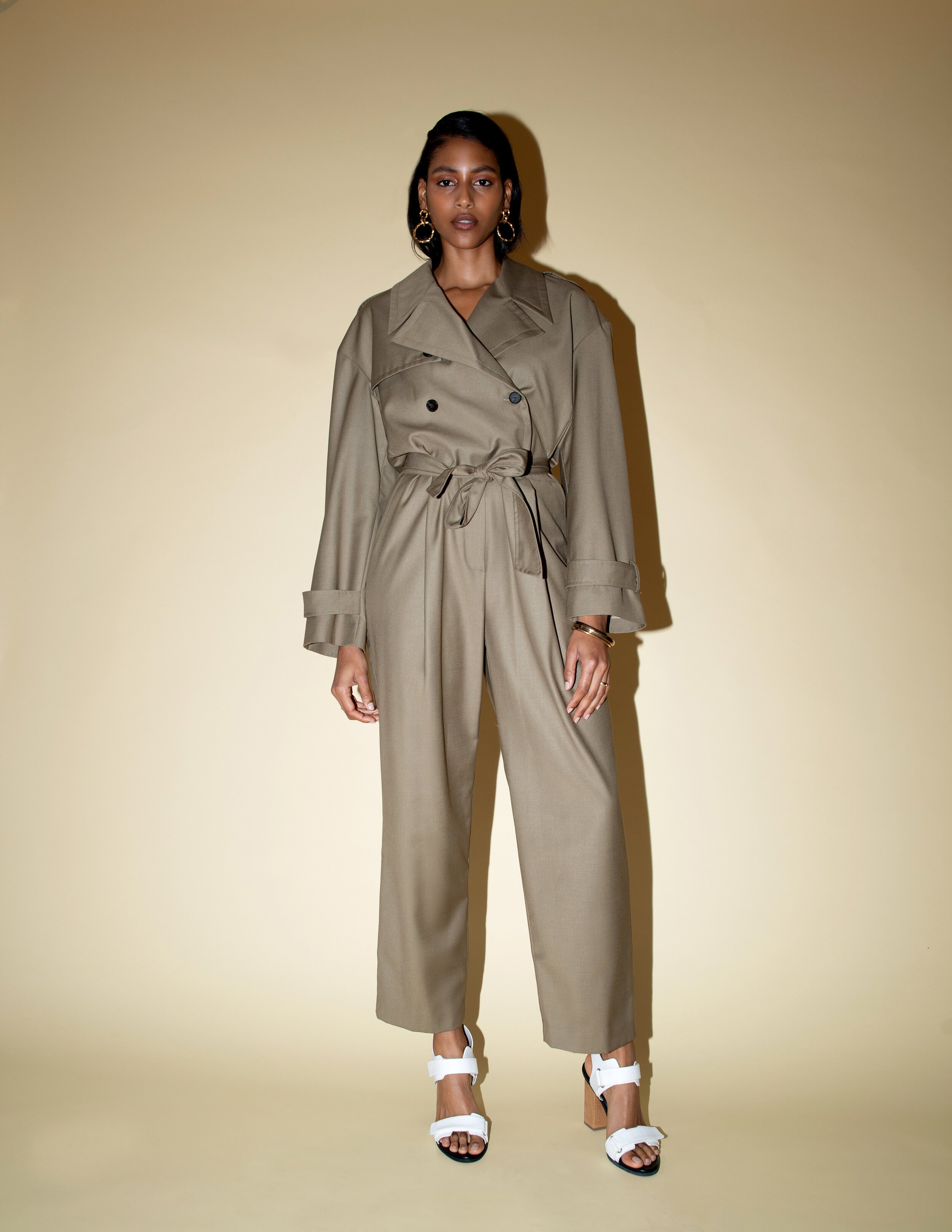 A full-length portrait of a fashionable adult woman in a tan trench coat and wide-leg pants, standing indoors against a pale background.