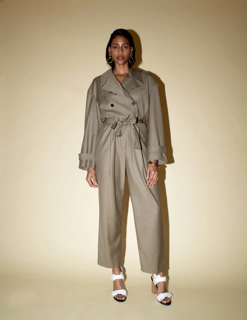 A full-length portrait of a fashionable adult woman in a tan trench coat and wide-leg pants, standing indoors against a pale background.