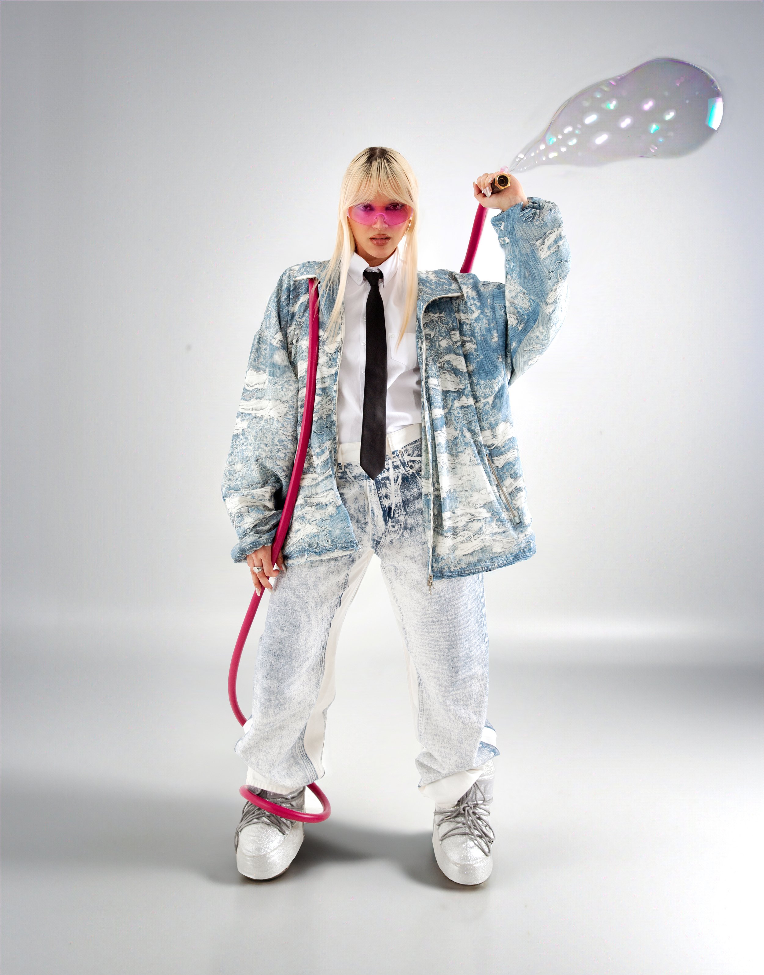 Full-length shot of a blonde woman in a denim jacket, pink glasses, and platform shoes, holding a pink hose with bubble effects.