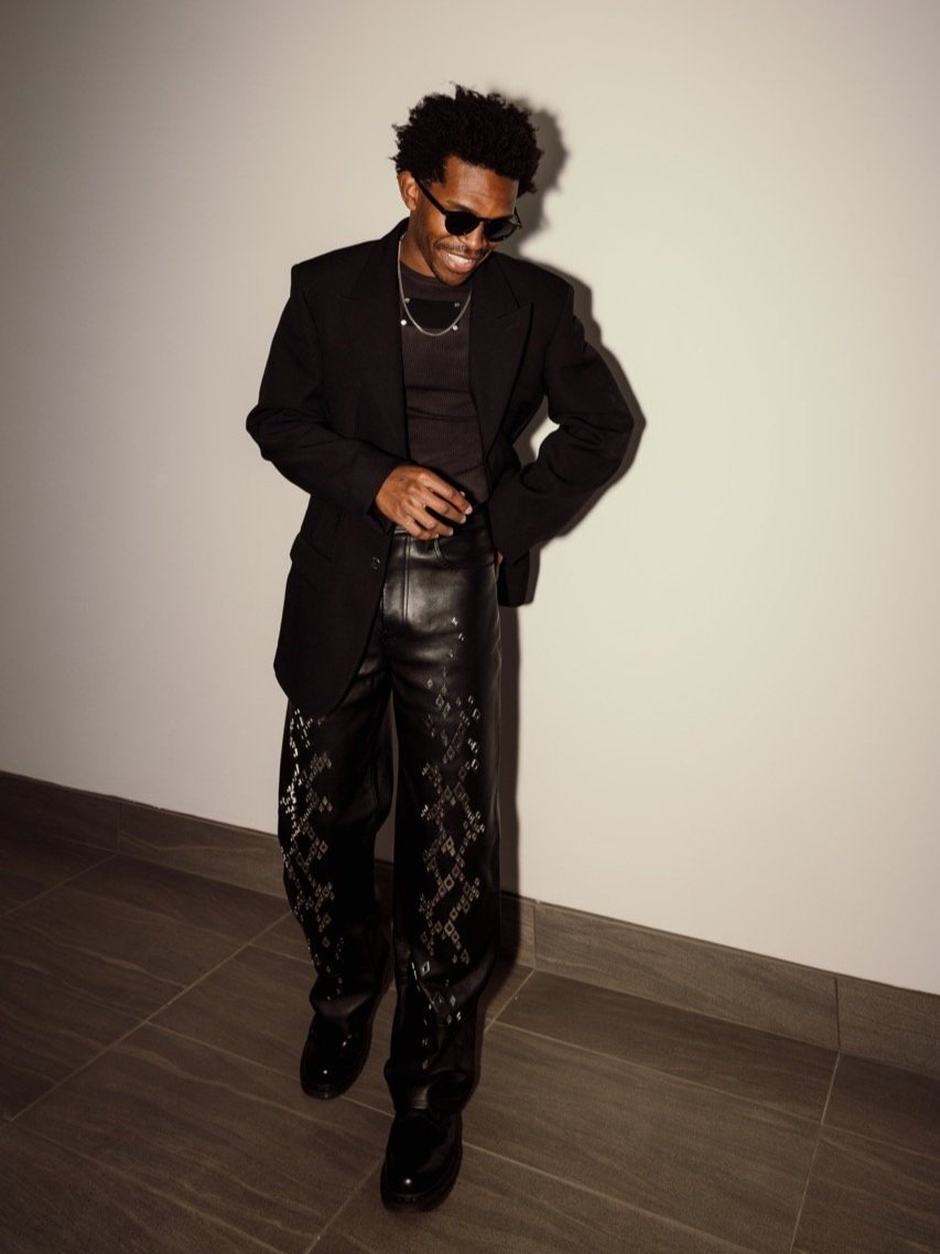 Young man poses in an indoor setting wearing a black blazer, black top, decorative leather pants, and black shoes, looking down and smiling.