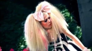 Woman with white hair, pale makeup, and a pink bow poses in a black and white dress against a blurred floral and greenery background.