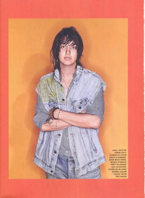 Portrait of a young adult male wearing a denim vest and jeans, standing against a bright orange background in a magazine editorial.