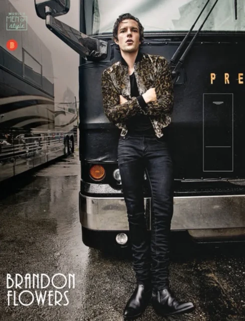 Brandon Flowers, lead singer of The Killers, stands by a black tour bus in a leopard-print jacket, black jeans and boots on a wet urban street.