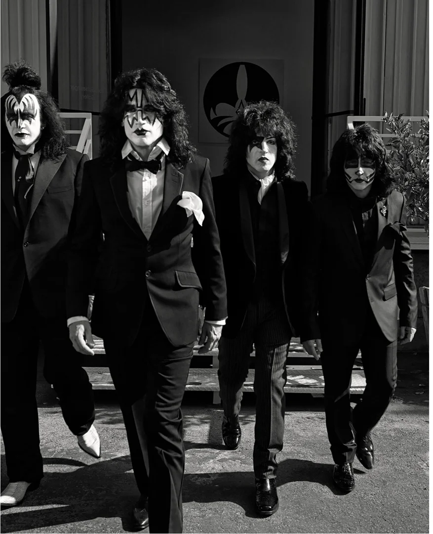 Four men in black suits with theatrical makeup walk toward the camera in front of a light-colored building with an emblem.