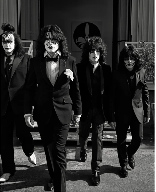 Four men in black suits with theatrical makeup walk toward the camera in front of a light-colored building with an emblem.