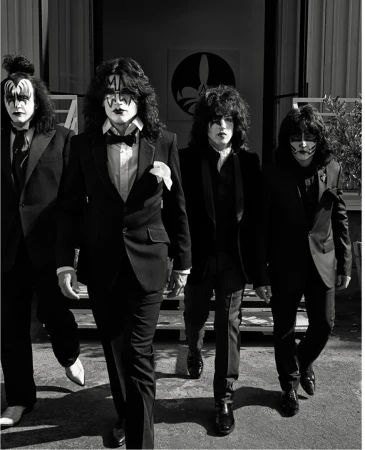 Four men in black suits with theatrical makeup walk toward the camera in front of a light-colored building with an emblem.