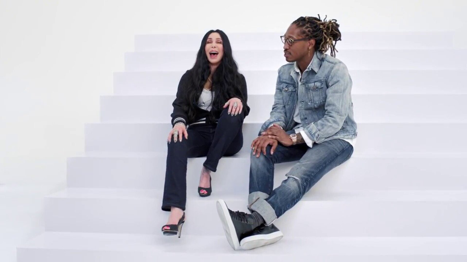 Cher and Future sit on white stairs. Woman wears black jacket, jeans, heels and man wears denim jacket, jeans, and black shoes.