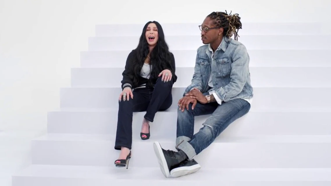 Cher and Future sit on white stairs. Woman wears black jacket, jeans, heels and man wears denim jacket, jeans, and black shoes.